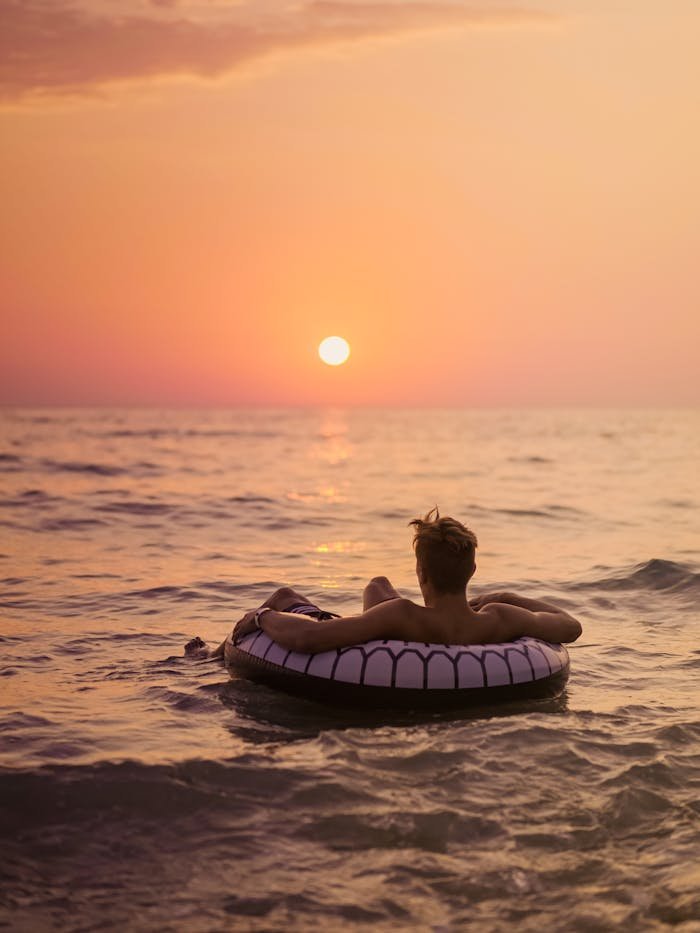 pexels-photo-4983184 Back view of unrecognizable male tourist lying in inflatable ring on wavy sea while contemplating bright sky with sun in evening